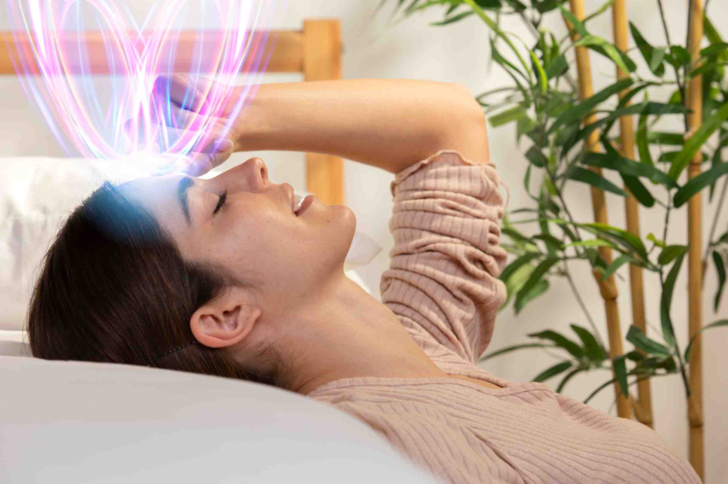 Woman lying on a bed with eyes closed, touching her forehead, as glowing purple and pink brain wave-like light patterns emerge from her forehead, symbolizing meditation, mental energy, or third eye activation; bamboo plants in the background.