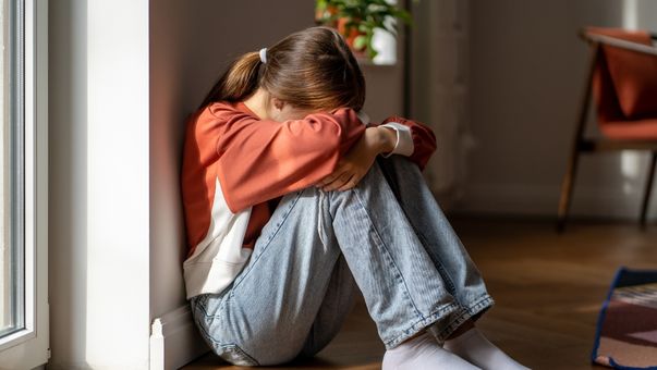 Young girl sitting on the floor by a window with her head down, hugging her knees, appearing sad or distressed in a quiet room.