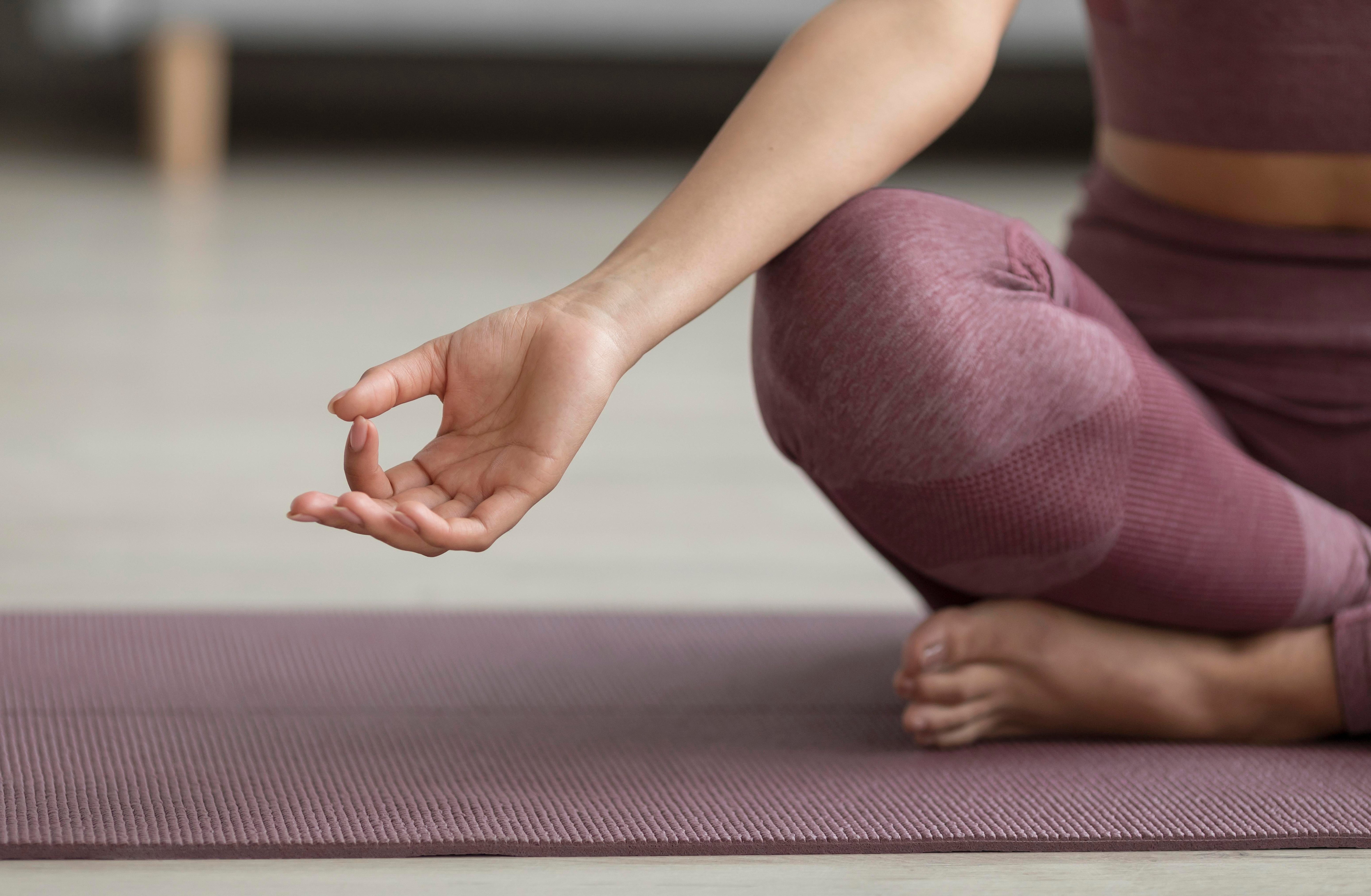 Person sitting cross-legged on a yoga mat in a meditative pose, with one hand resting on the knee in a Gyan mudra (thumb and index finger touching), wearing pink athletic wear.